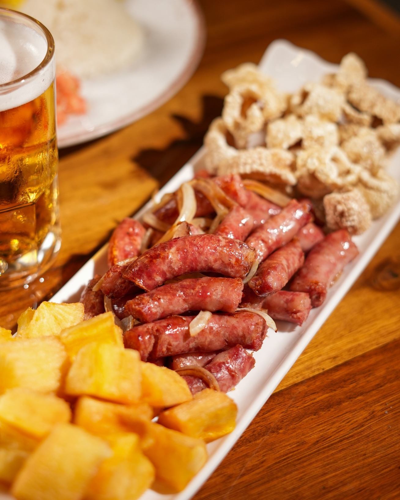 Travessa de linguiça acebolada, torresmo e mandioca frita ao lado de um copo de chopp