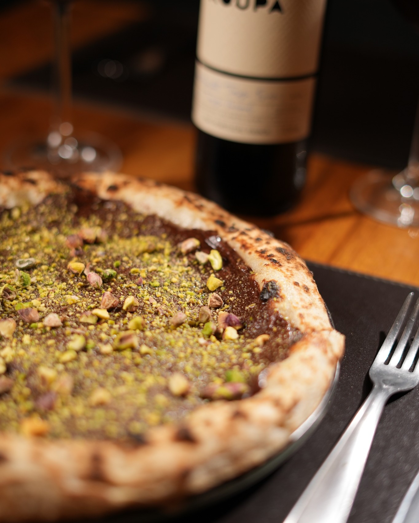 Pizza de pistache saída do forno, com borda tostada, ao lado de uma garrafa de vinho